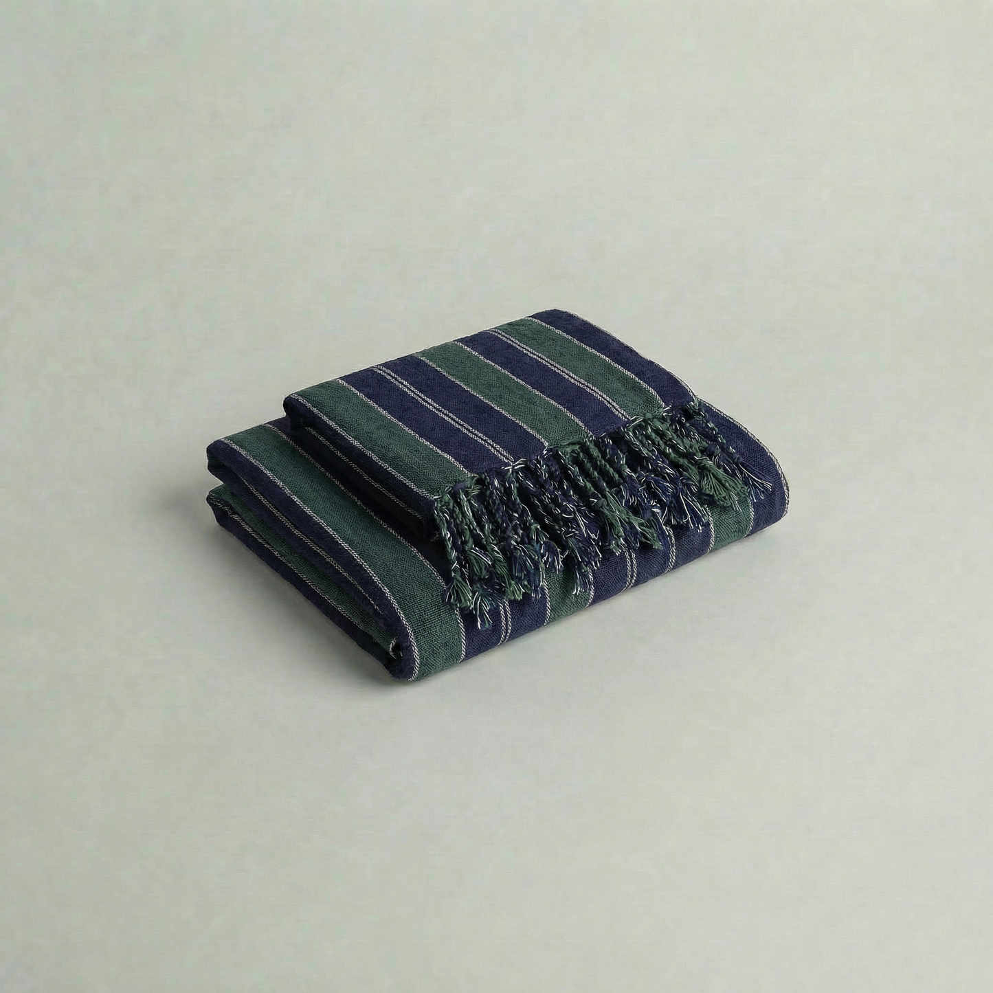 Folded handwoven atelier orea cotton towel in dark green and navy with fringes on a light gray background