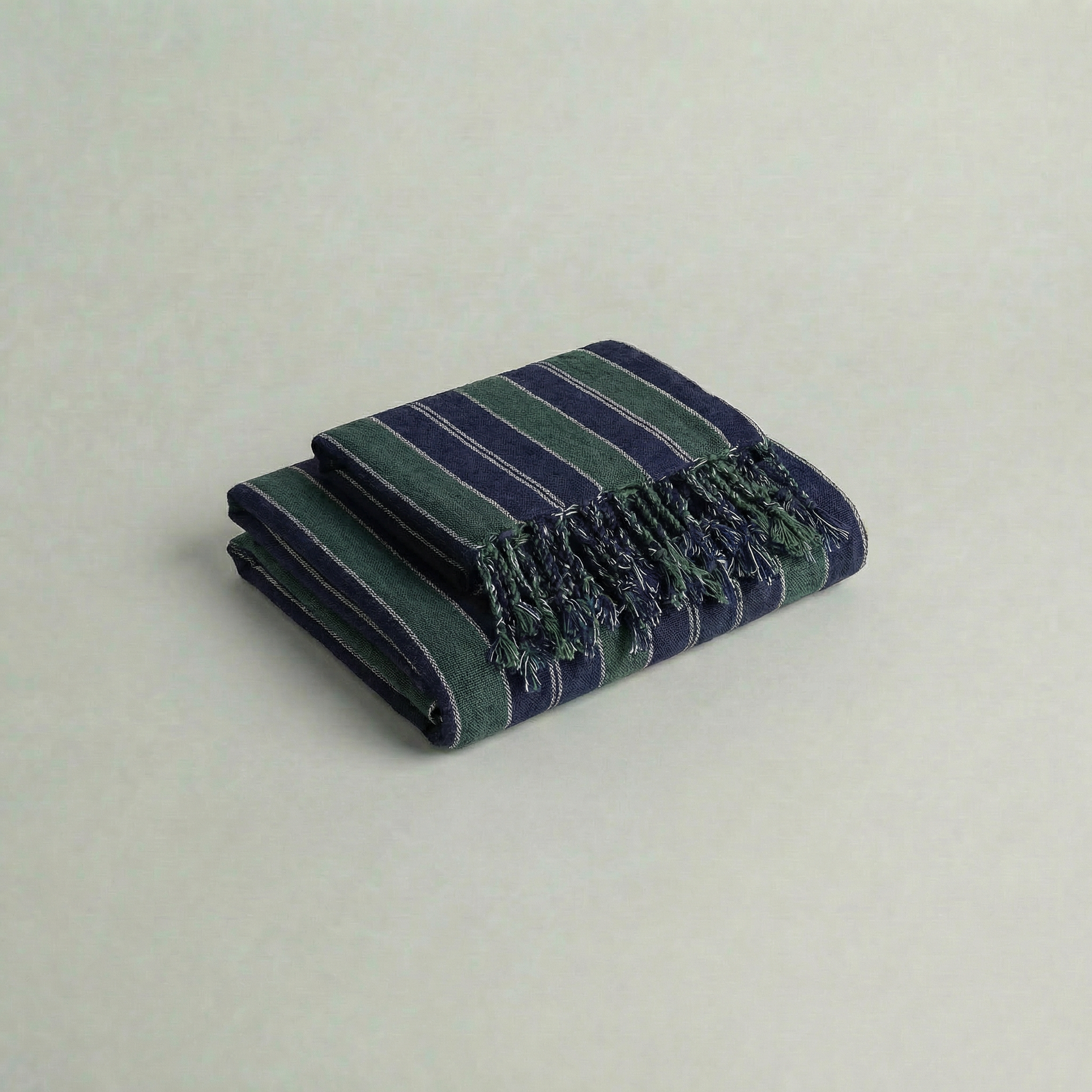 Folded handwoven atelier orea cotton towel in dark green and navy with fringes on a light gray background