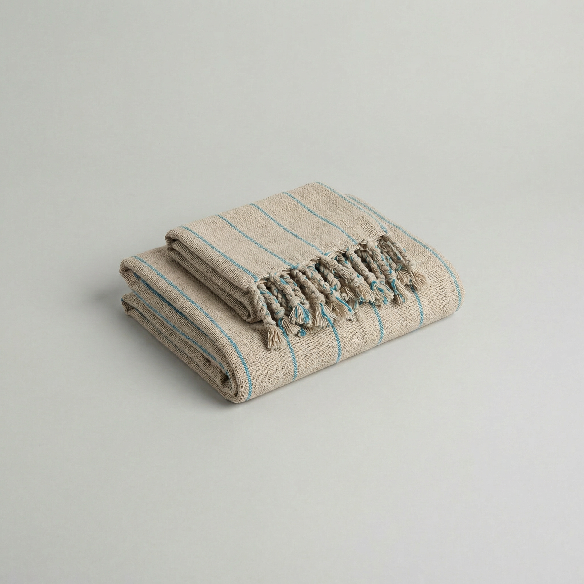 Folded handwoven slub cotton atelier orea beige towel with blue stripes and tassels on a light gray background