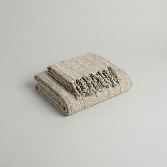 Folded handwoven slub cotton atelier orea beige towel with blue stripes and tassels on a light gray background