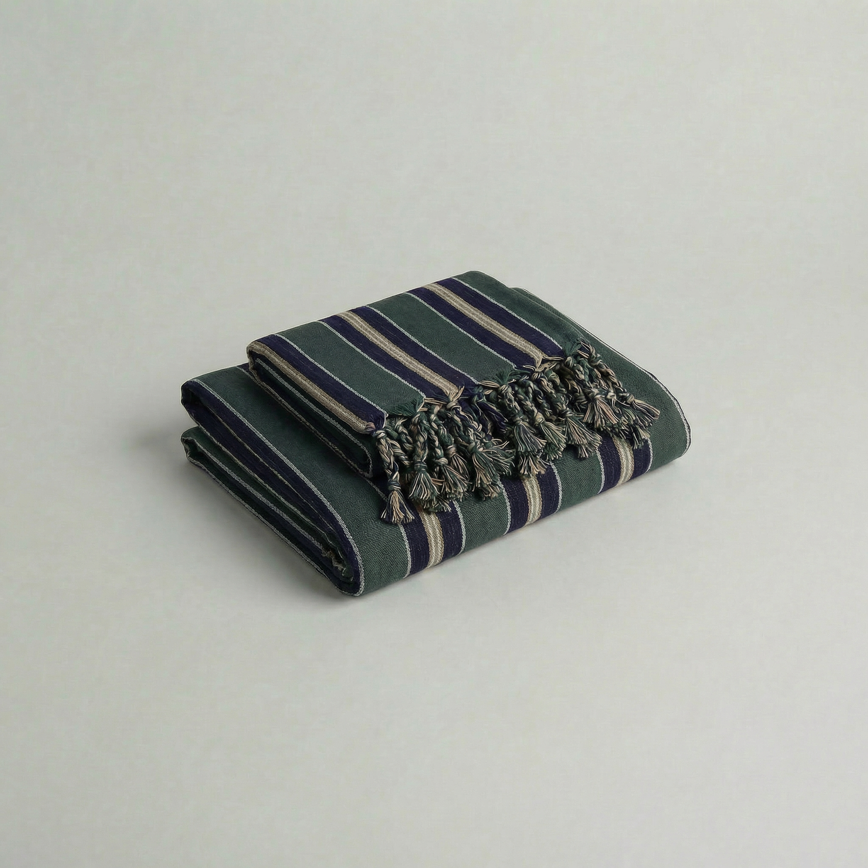 Folded handwoven atelier orea cotton towel in navy, dark green stripes with tassels on a light gray background