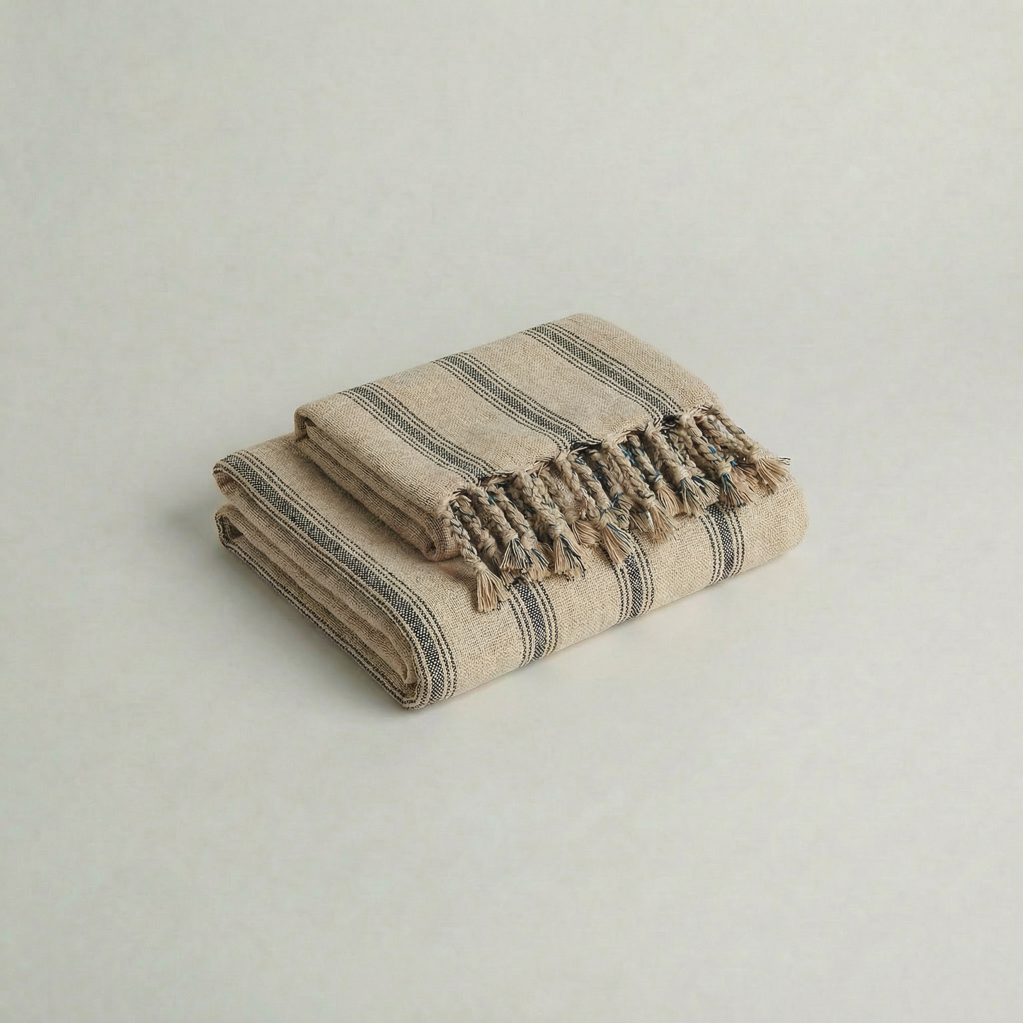 Folded handwoven slub cotton atelier orea beige and gray striped towel with tassels on a light gray background