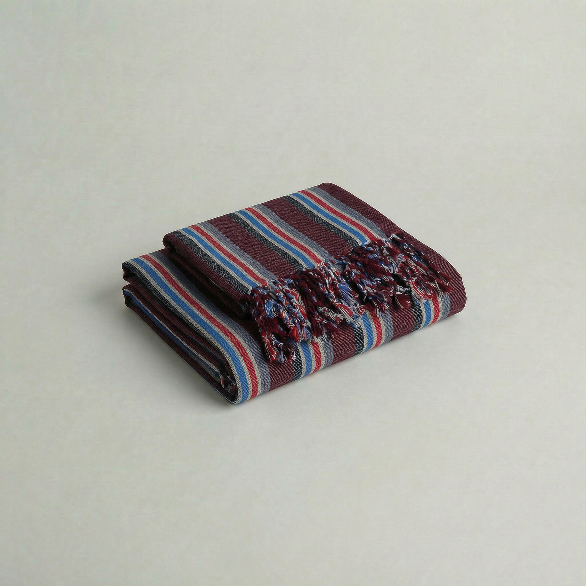 Folded handwoven cotton atelier orea maroon, blue, red striped towel with frayed edges on a light gray background