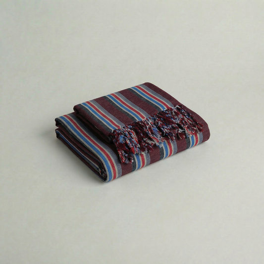 Folded handwoven cotton atelier orea maroon, blue, red striped towel with frayed edges on a light gray background