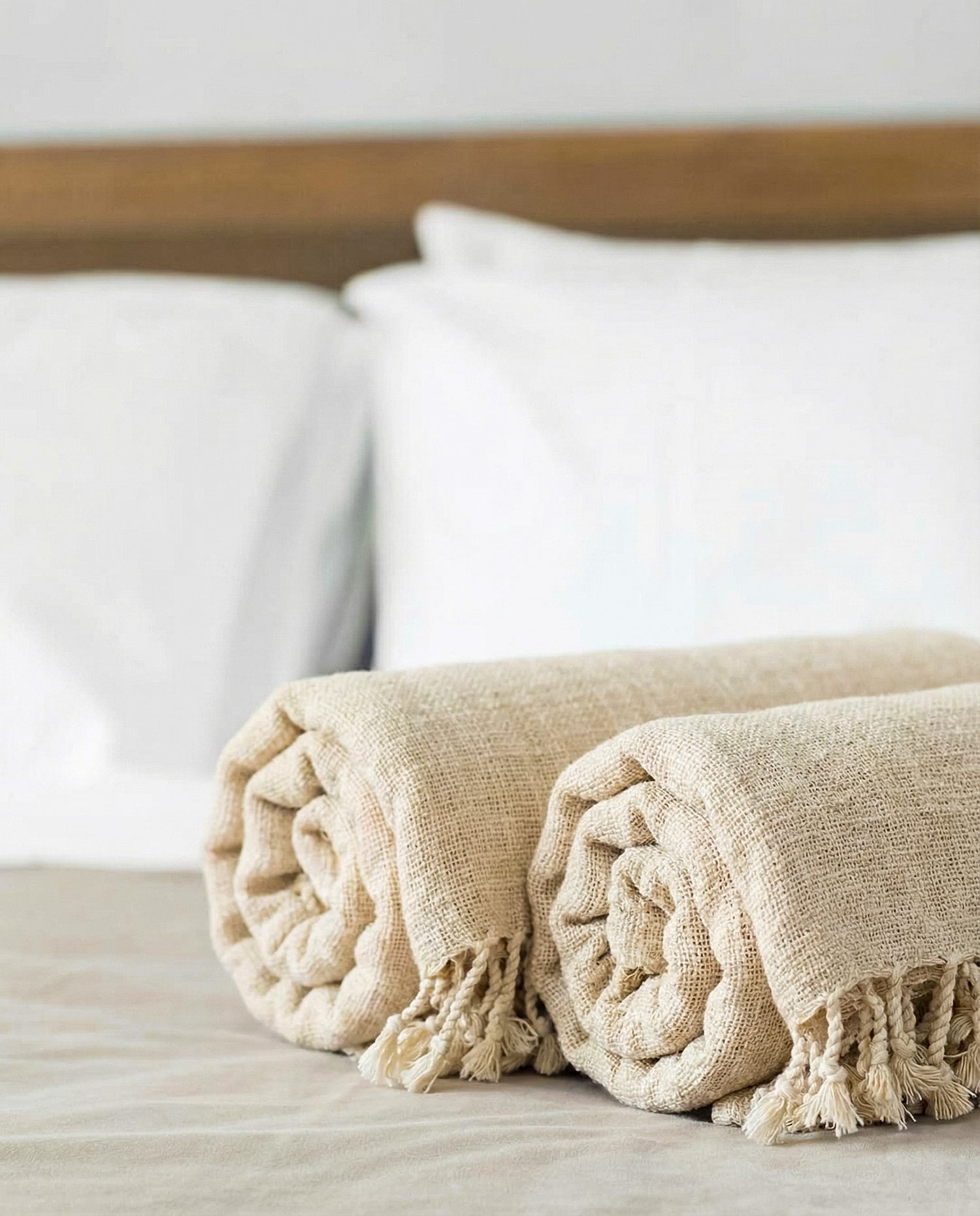 Two beige folded handwoven slub turkish cotton towels on a bed with white pillows and a wooden headboard.
