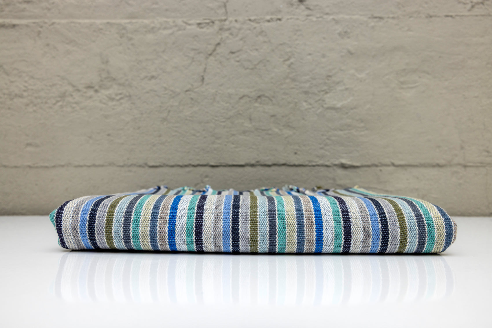Folded atelier orea handwoven turkish cotton towel with blue, turquoise, cyan and navy stripes on a white surface and gray background.