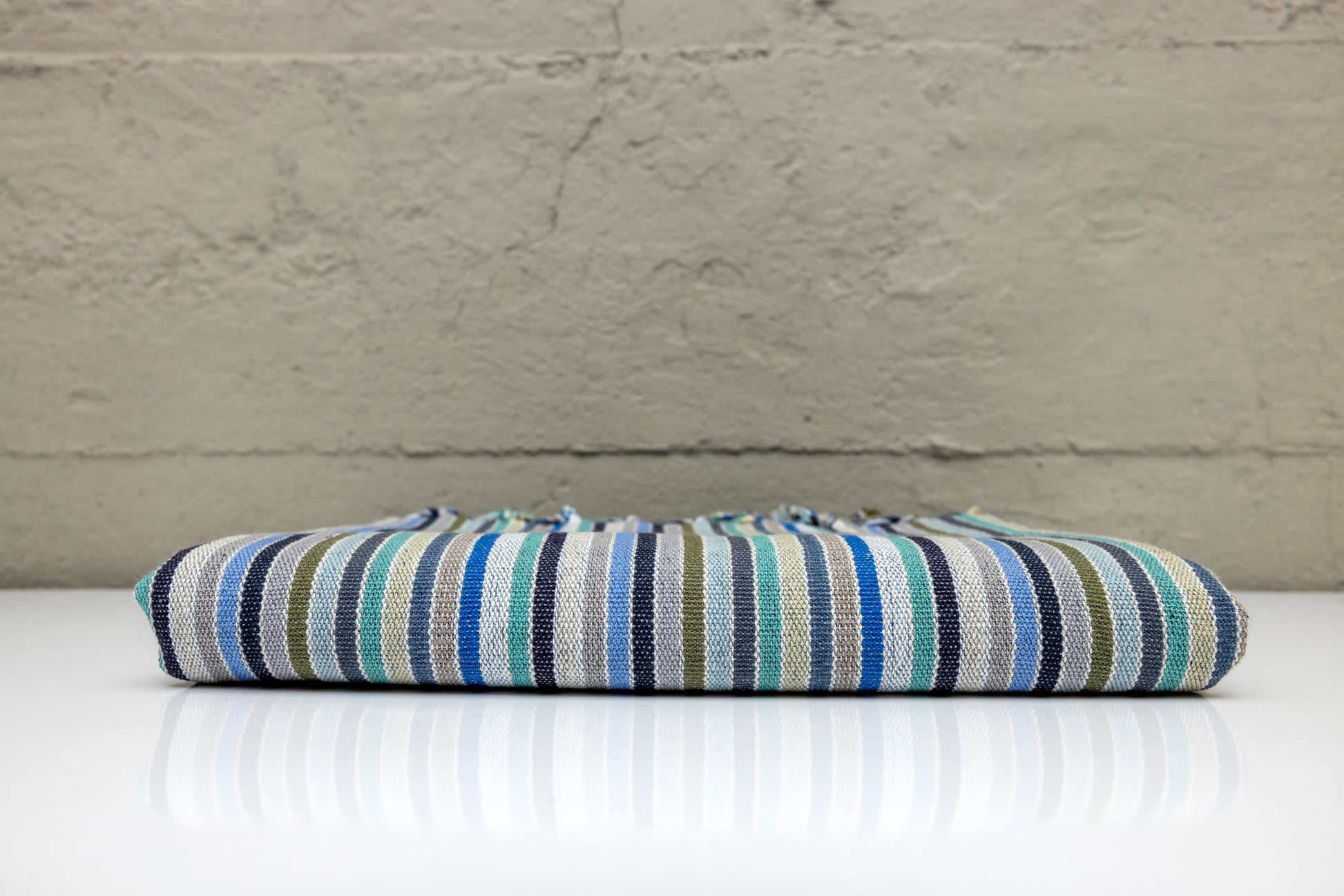 Folded atelier orea handwoven turkish cotton towel with blue, turquoise, cyan and navy stripes on a white surface and gray background.