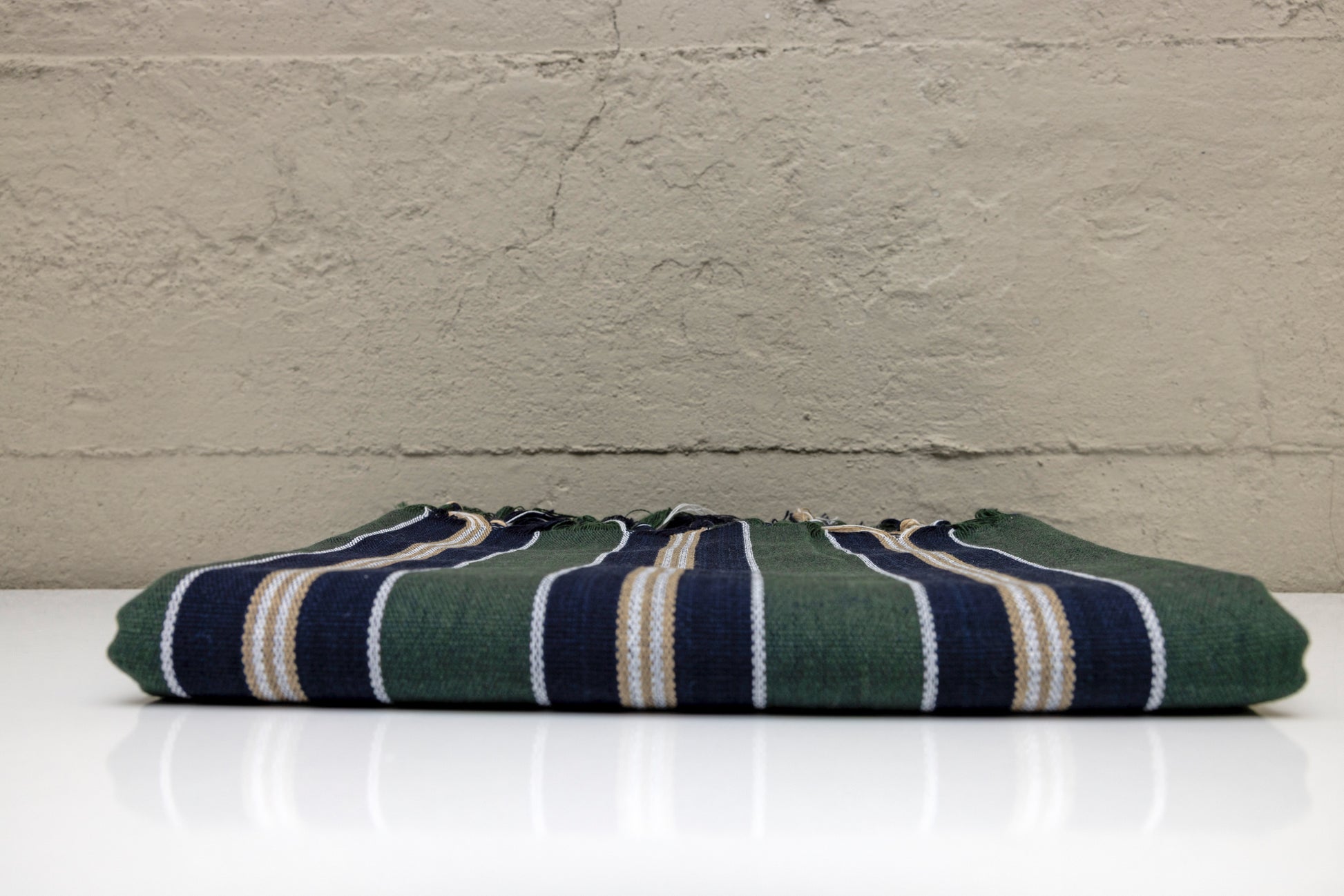 Folded atelier orea handwoven turkish cotton towel with dark green navy and beige stripes with a white surface with a gray textured wall background