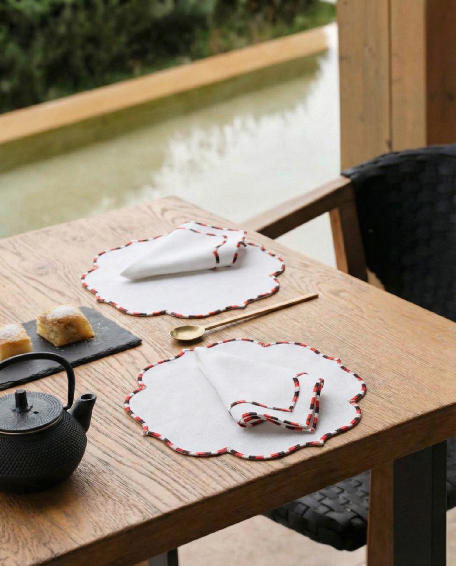 Table setting with white embroidered placemats and napkins on a wooden table by a window.